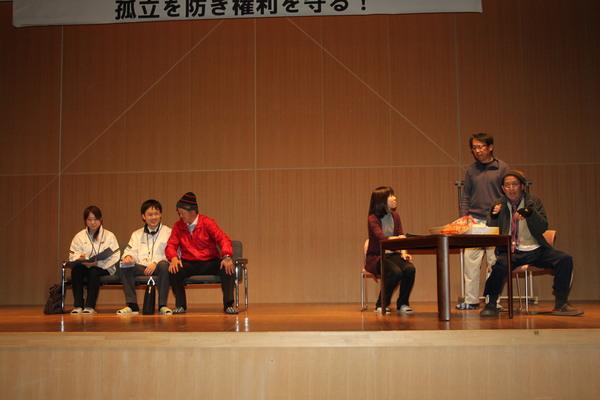 舞台の上でソファーに腰かけて話をしている男女3名とテーブルに座っている女性と話をしている男性が2名写っている写真